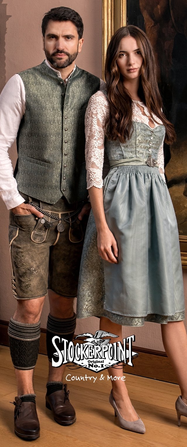 Hombre y mujer de pie en el interior vistiendo ropa tradicional bávara, que incluye lederhosen, chaleco, blusa de encaje, vestido dirndl y tacones.