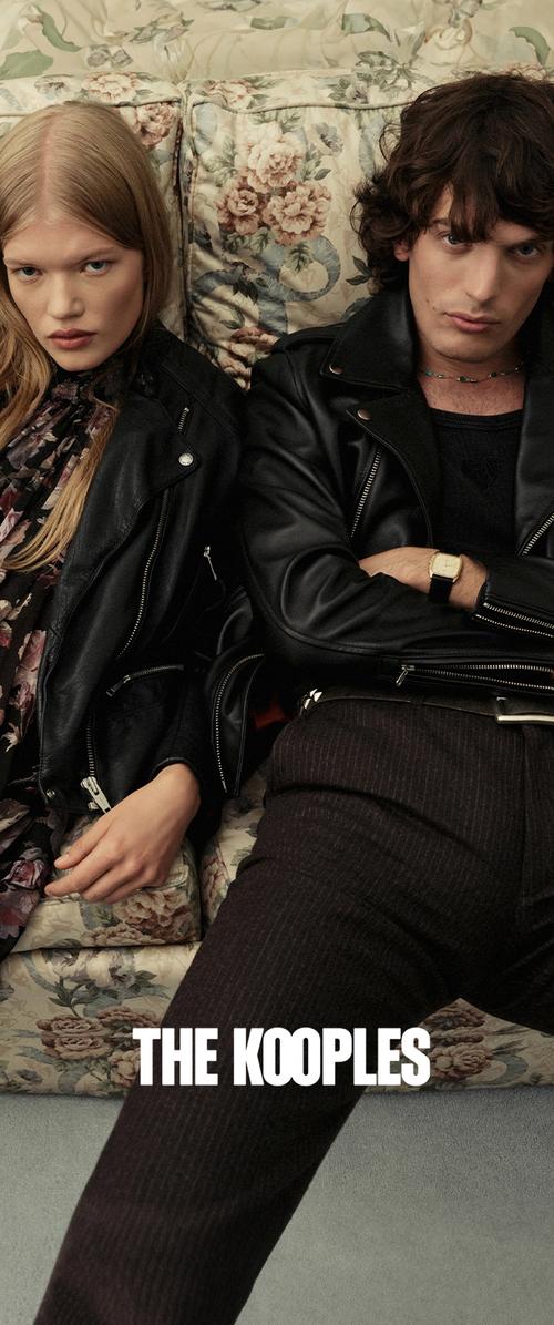Deux mannequins sont assis sur un canapé à motifs floraux, portant des vestes en cuir noires. L'arrière-plan présente un tapis texturé de couleur neutre.