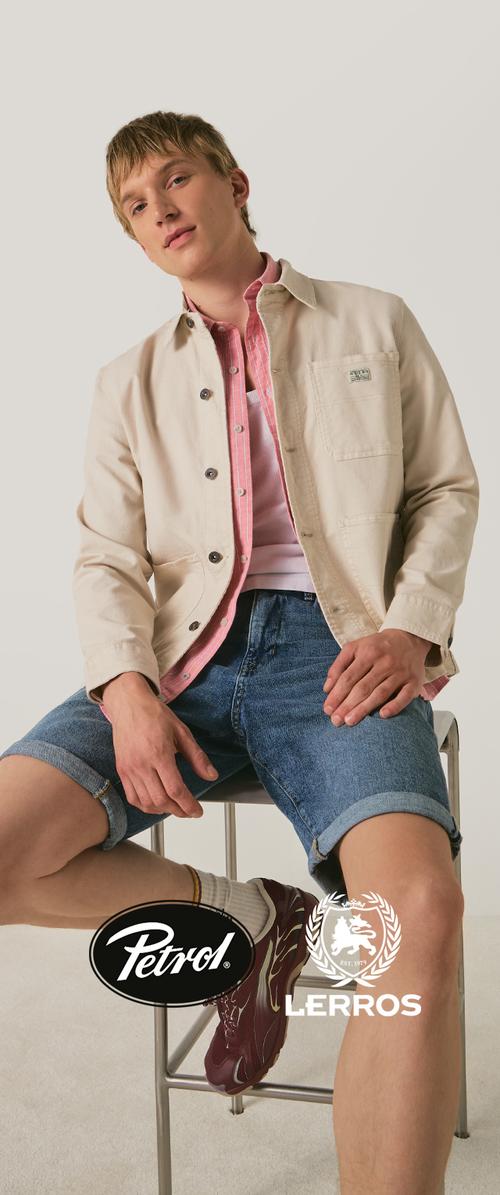 Jonge man zit op een kruk, draagt een beige jas, roze overhemd, wit hemd, spijkershort, witte sokken en rode sneakers.
