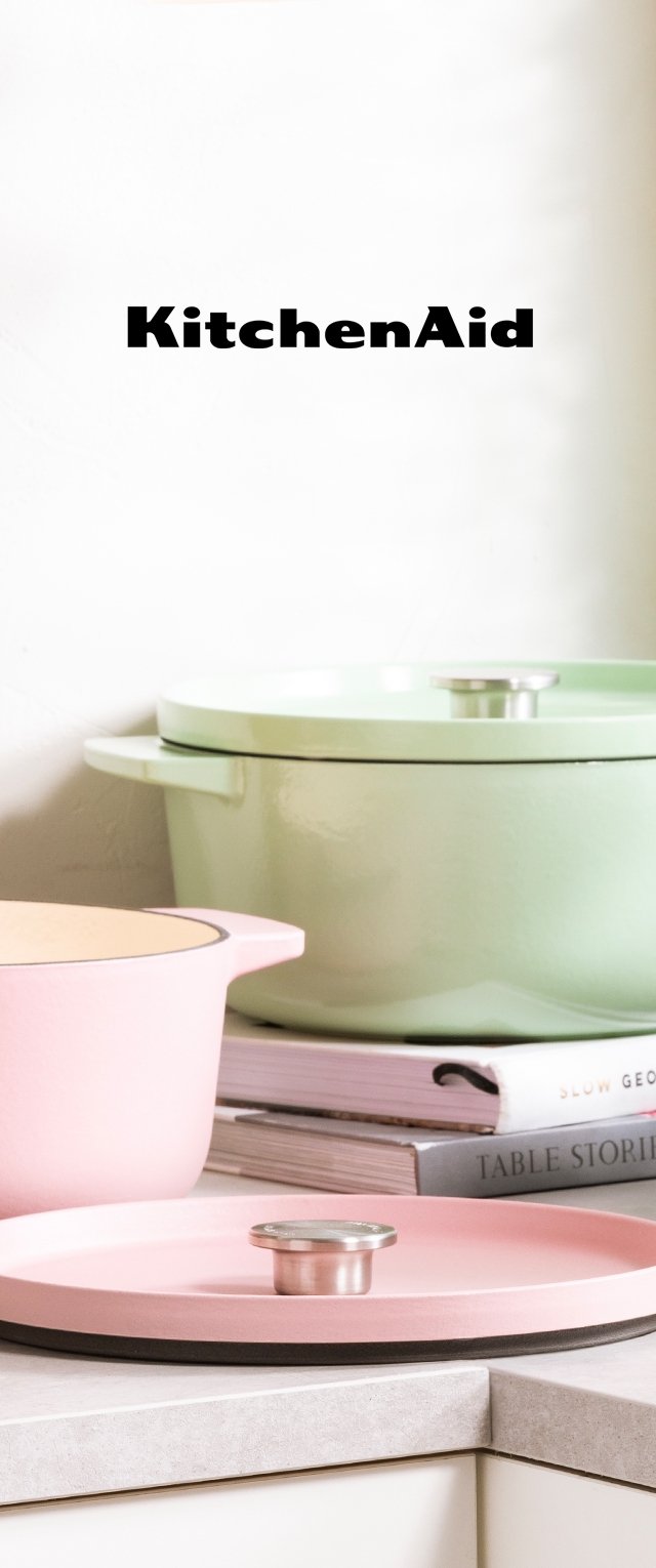 Drei Kochgeschirrteile von KitchenAid: ein pinkfarbener runder Topf mit einem Metallgriff, ein grüner Deckel mit einem silbernen Knopf und ein pinkfarbener Tablett mit einer schwarzen Kante.