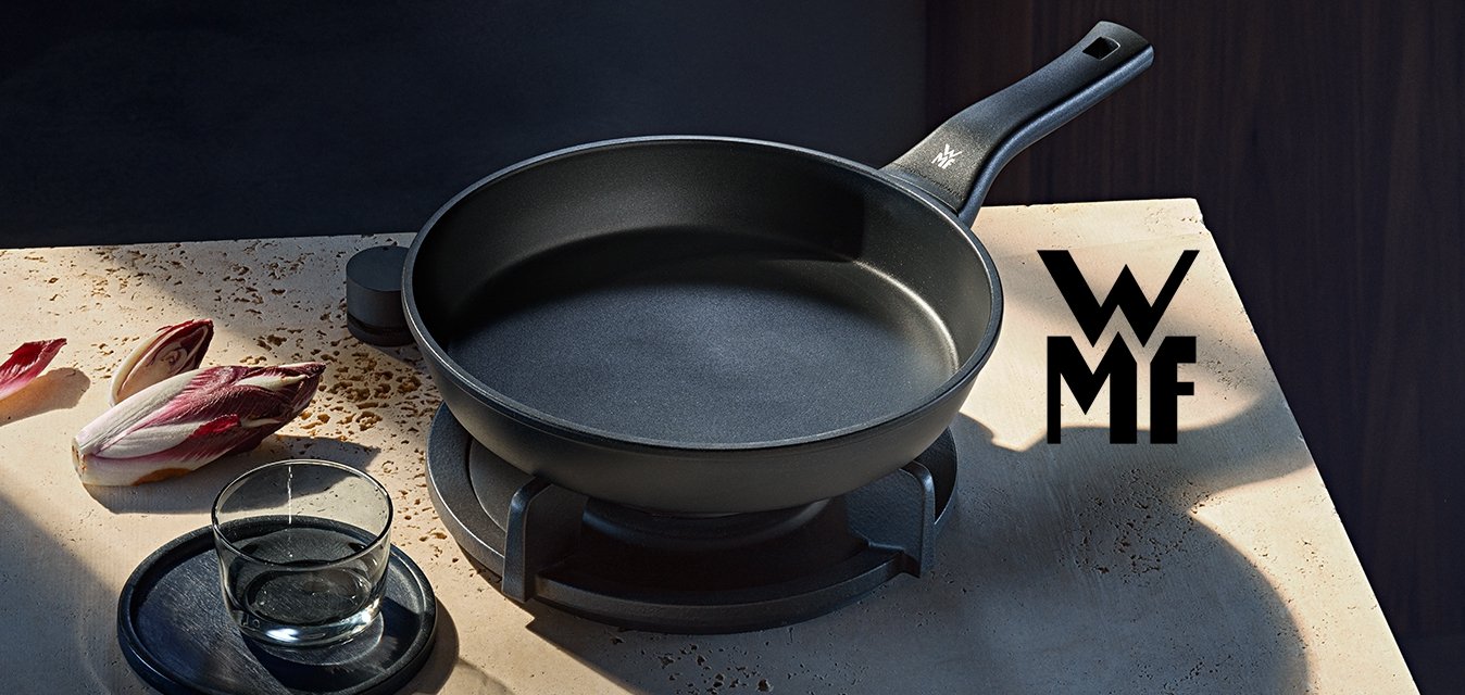 Černá pánev na sporáku s nasekanými listy čekanky, sklenice vody na podtácku a černý talíř na kamenné pracovní desce.