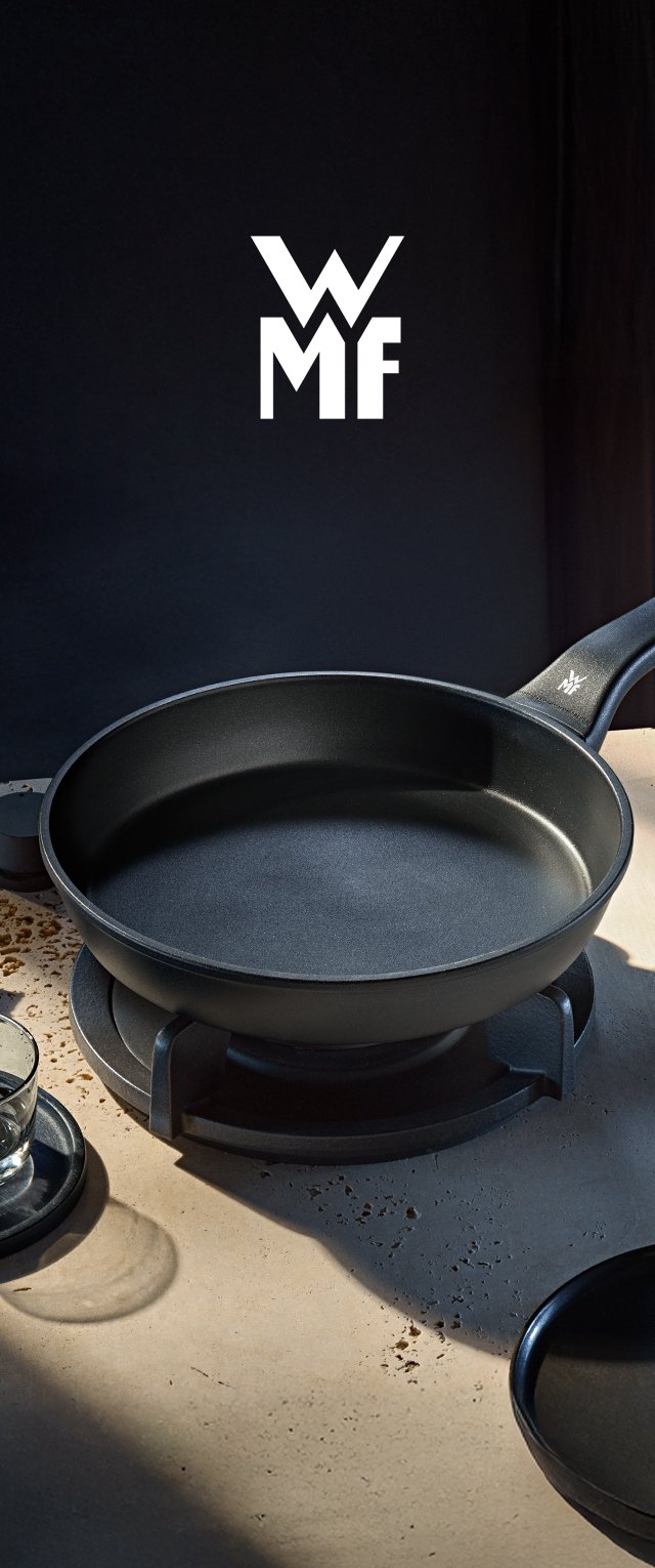 Sartén negra WMF en la estufa con mango, colocada sobre una encimera beige moteada, con un vaso sobre un posavasos cerca.