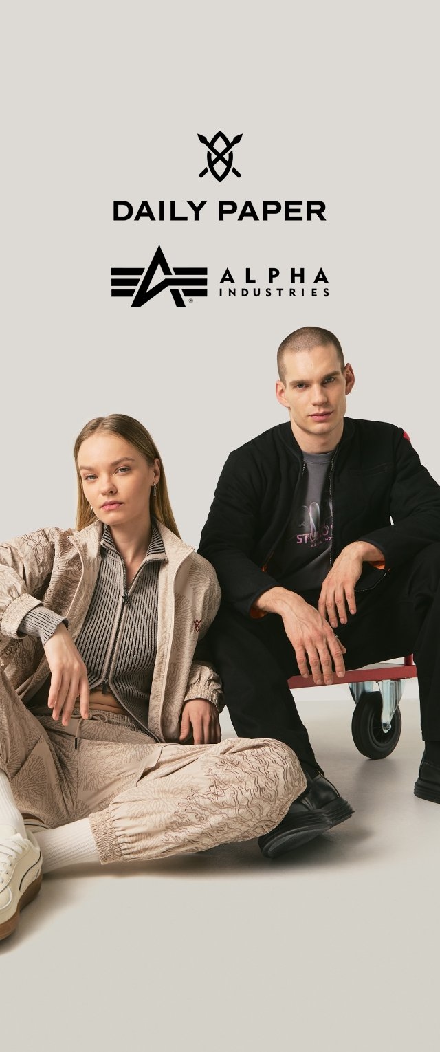 Zwei Modelle sitzen auf dem Boden, Frau in beigem, gemustertem Trainingsanzug und Mann in schwarzer Jacke und Hose, mit den Logos von Daily Paper und Alpha Industries darüber.