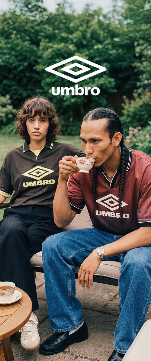 Deux personnes portant des t-shirts Umbro sont assises en plein air ; l'une boit du thé dans une tasse fleurie tandis que l'autre regarde devant, avec de la verdure en arrière-plan.