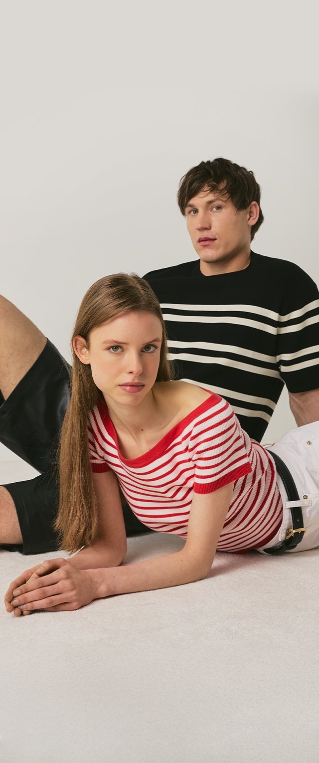 Nuori nainen punavalkoraidallisessa paidassa makaa vatsallaan lattialla, nuori mies mustavalkoraidallisessa paidassa istuu hänen takanaan yksiväristä taustaa vasten.