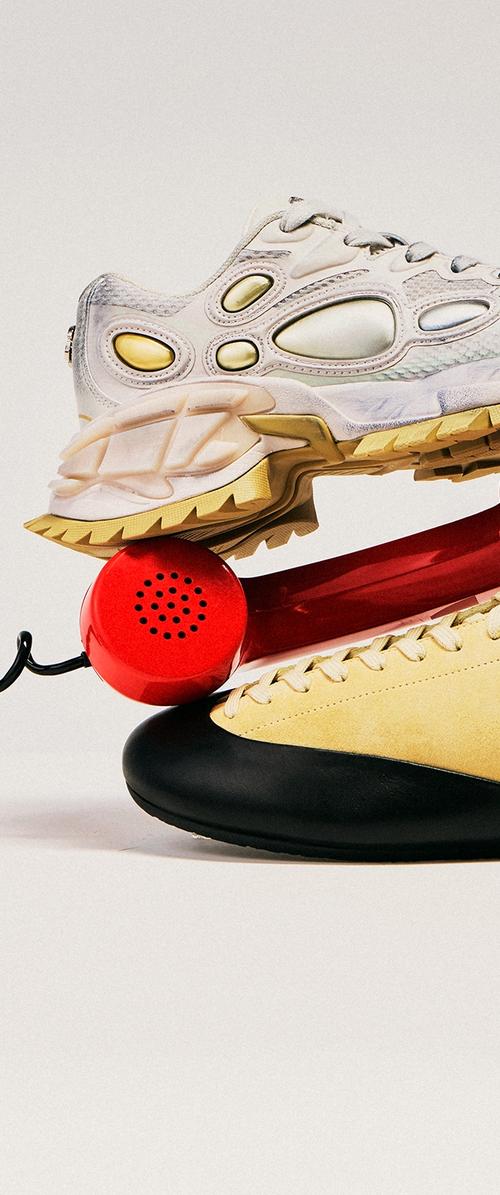 Baskets blanches et beiges empilées sur une chaussure jaune et noire, avec un combiné de téléphone rétro rouge équilibré entre elles.