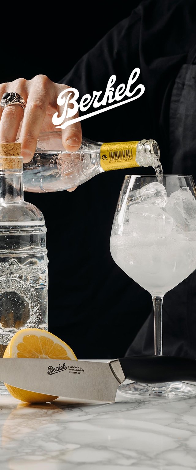 Un bicchiere con ghiaccio è riempito con un liquido trasparente versato da una bottiglia di vetro. Un coltello d'argento giace su una superficie di marmo vicino a una fetta di limone.