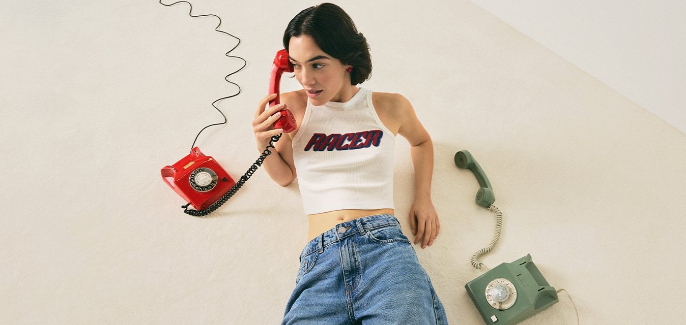 Žena v bílém crop topu a džínách leží na podlaze, drží červený otočný telefon, vedle ní leží zelený otočný telefon.