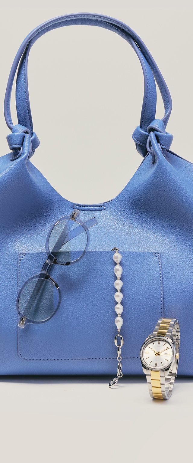 Blå läderhandväska med knutna handtag, genomskinliga runda solglasögon upphängda på fickan, pärlarmband och silver-guld armbandsur på sidan.