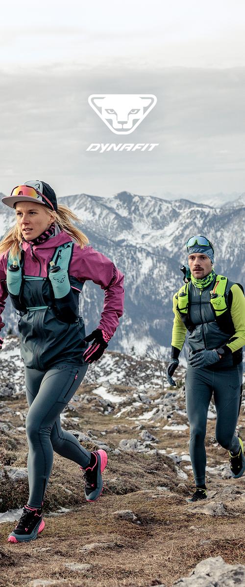Zwei Wanderer in Winterausrüstung laufen auf einem felsigen Bergweg mit schneebedeckten Gipfeln im Hintergrund und dem Dynafit-Logo darüber.