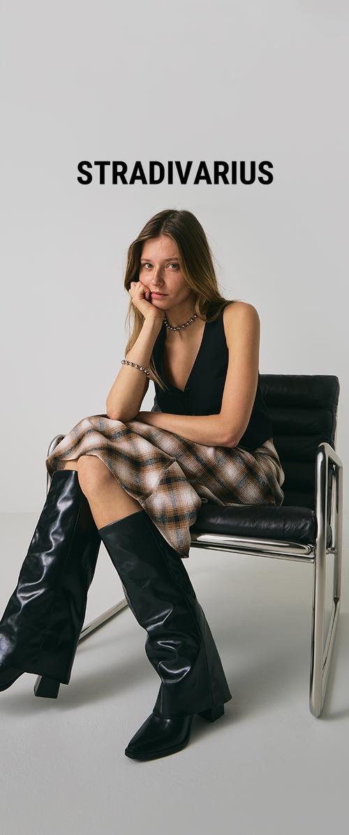Jonge vrouw met lang haar zit op een zwarte leren stoel, gekleed in een zwart mouwloos topje, een geruite rok, kniehoge zwarte laarzen en zilveren sieraden.