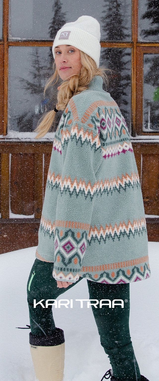 Poncho in fleece verde chiaro con motivi geometrici colorati, indossato su pantaloni verdi scuri e stivaletti color crema, completato da un berretto in maglia bianco.