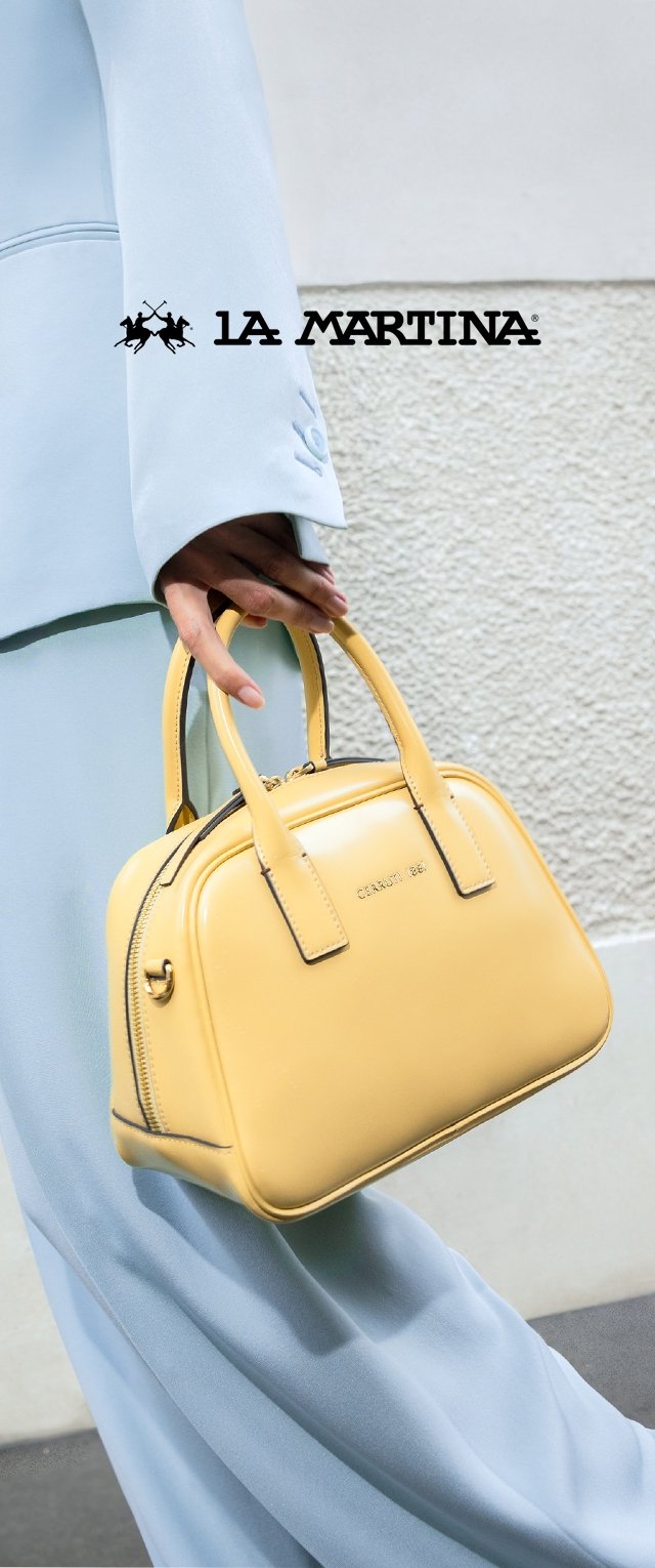 Żółta skórzana torebka o zaokrąglonym kształcie, z podwójnymi uchwytami u góry i czarnymi szwami. Na tle jasnoniebieskiego materiałowego pokrowca.