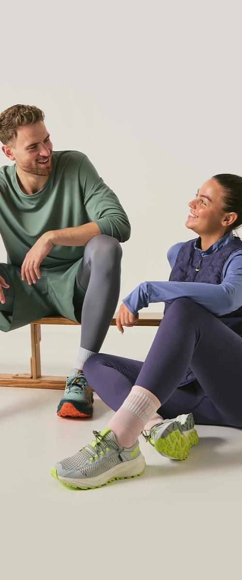 Mann und Frau in Sportkleidung, die sitzen und lächeln, während sie sich während des Trainings gegen einen einfachen hellen Hintergrund ausruhen.