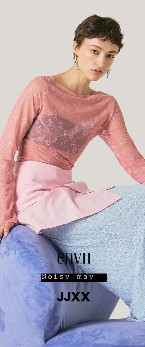 Joven mujer con cabello corto y oscuro que lleva una blusa rosa transparente con estampado floral, falda rosa claro y pantalones texturizados azul claro, sentada en una silla azul.
