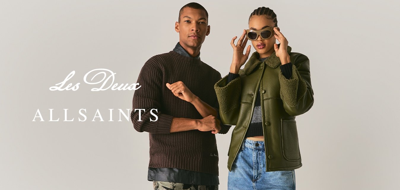 Dva lidé stojí proti jednoduchému pozadí; jeden má na sobě tmavou svetr a vzorované kalhoty, druhý olivovou bundu, džíny a sluneční brýle.