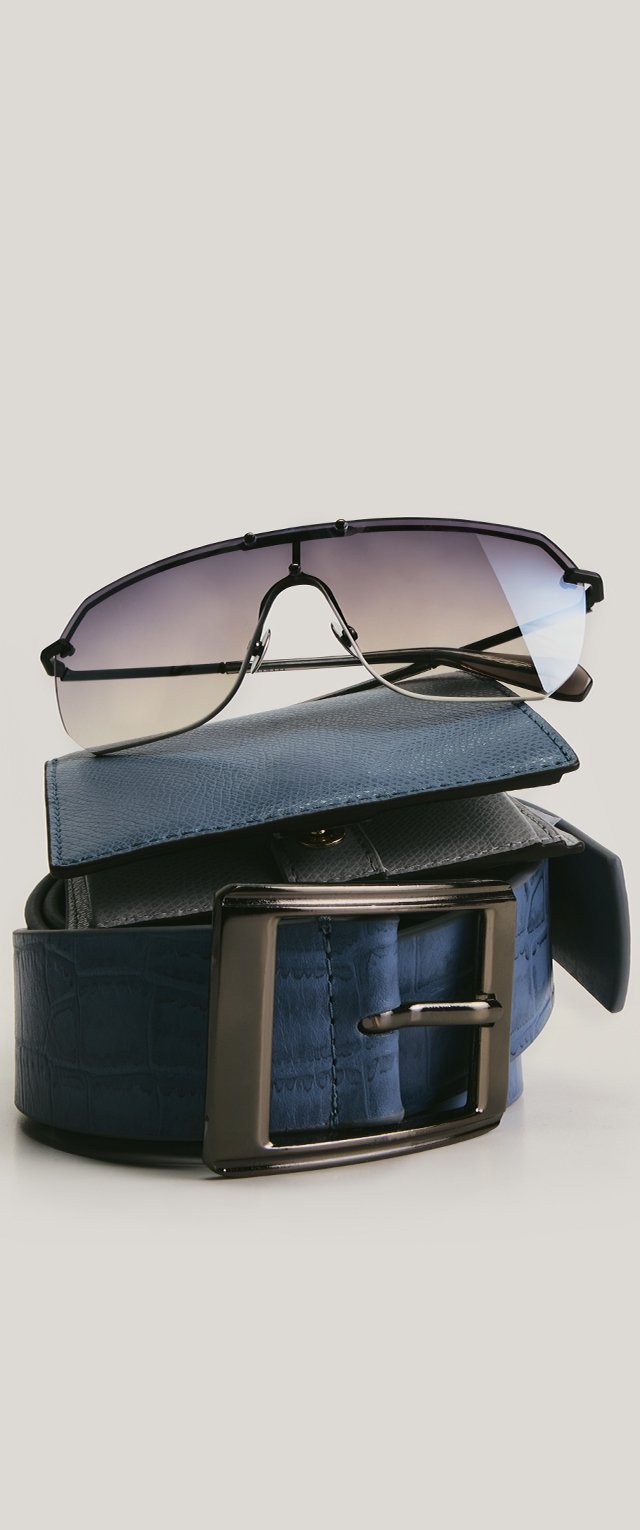 Cintura in pelle blu texturizzata con fibbia in metallo, portafoglio pieghevole e occhiali da sole aviator con lenti sfumate poggiati sopra.