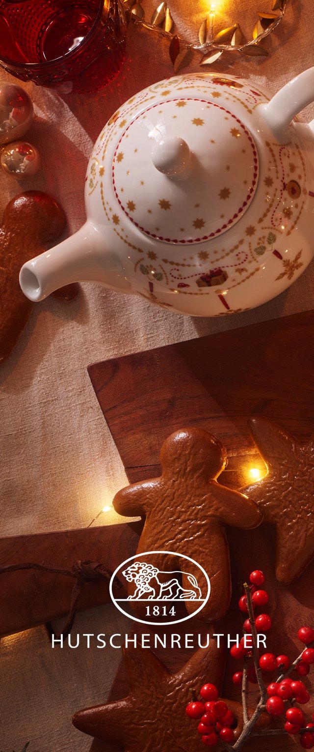 Tetera de porcelana blanca con un patrón de estrellas doradas y rojas, acompañada de galletas de jengibre y decoración festiva roja sobre un fondo de tela texturizada.