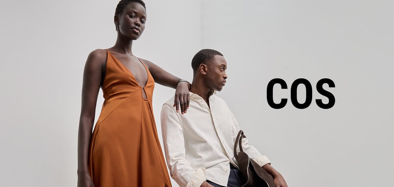 Dlouhé oranžové šaty s hlubokým výstřihem a bočním rozparkem, v kombinaci s lehkou texturovanou košilí a tmavými kalhotami, doplněné hnědou kabelkou.