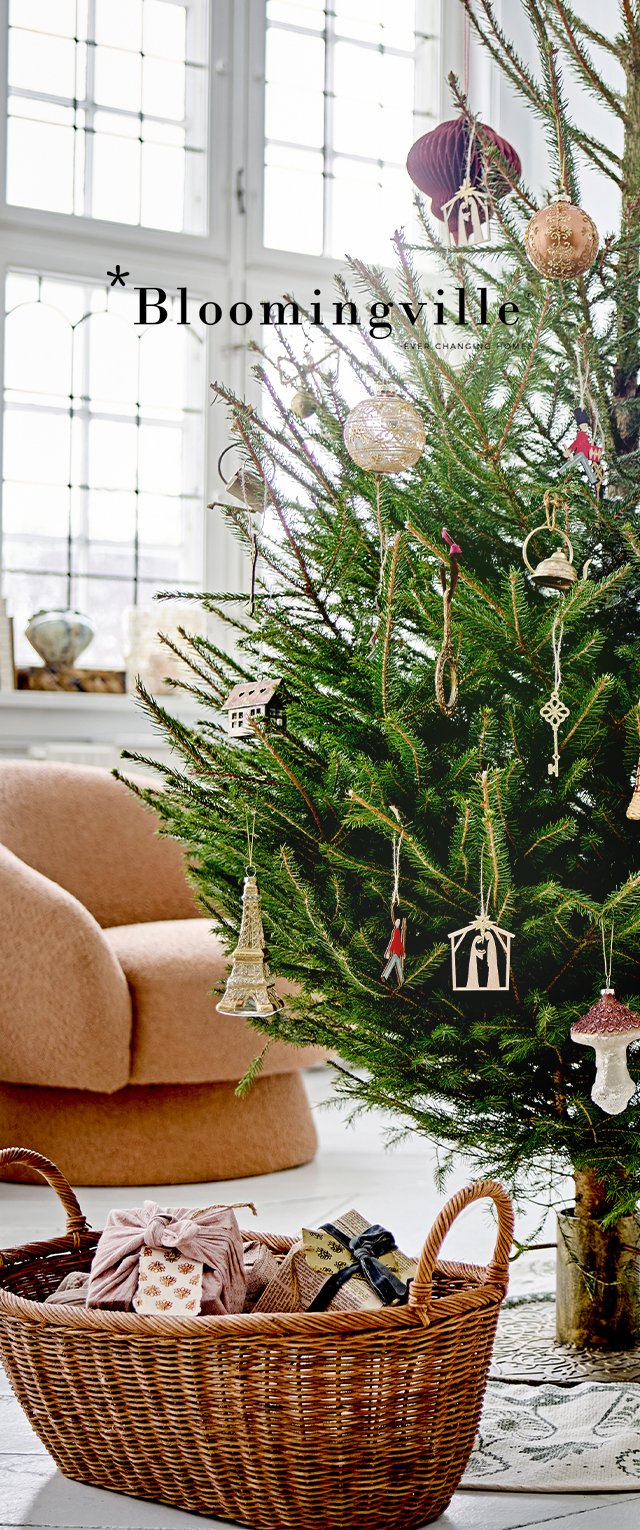 Albero di Natale con aghi verdi, decorato con vari ornamenti, accanto a un cesto intrecciato contenente regali incartati e una morbida poltrona rosa.