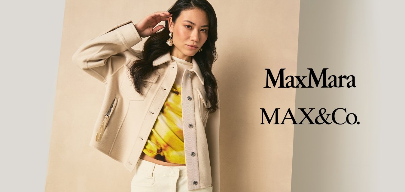 Žena s dlouhými tmavými vlasy, oblečená v béžovém saku, žlutém topu s vzorem a světlých kalhotách, stojí před neutrálním pozadím.