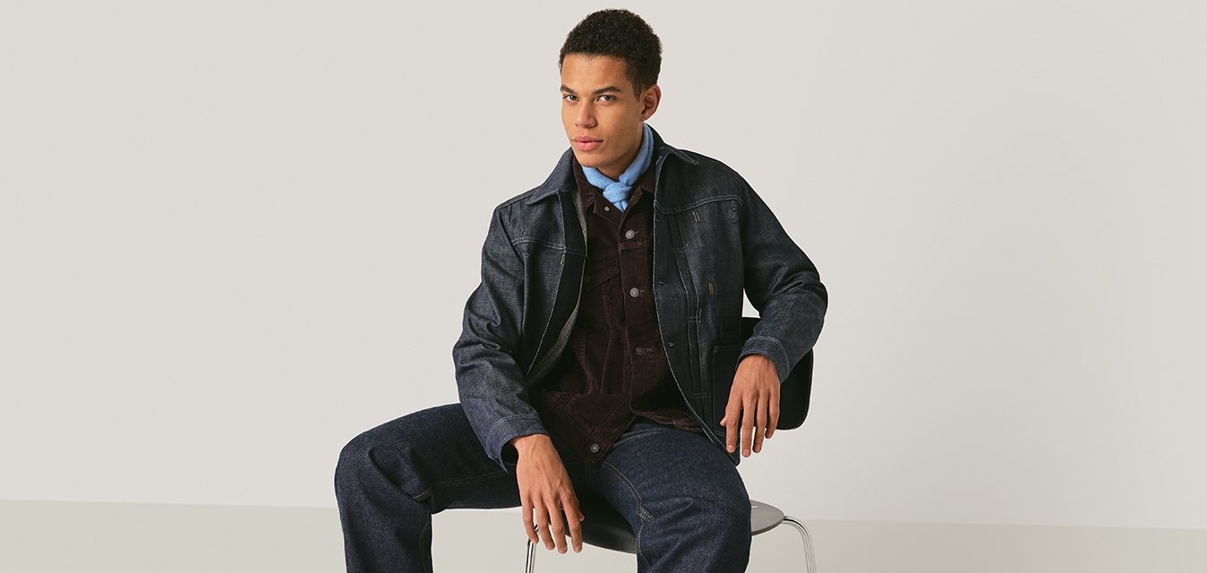 Mladý muž oblečený v tmavé texturované bundě, modré košili, manšestrové vestě, džínových kalhotách a černých botách sedí na židli před jednobarevným pozadím.