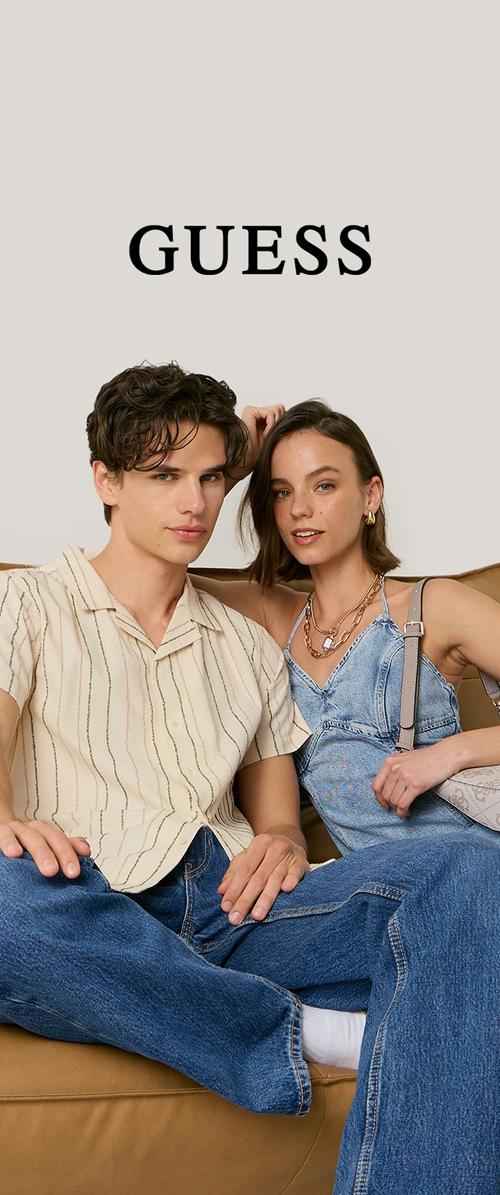 Joven hombre y mujer sentados en un sofá, vestidos con conjuntos veraniegos casuales con denim, posando bajo el logo de la marca "GUESS".