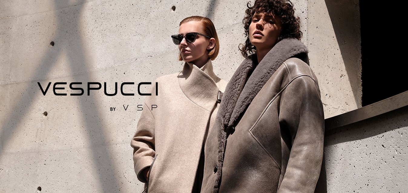 Dvě ženy v módních nadměrných zimních kabátech stojí opřené o texturovanou betonovou zeď, na které stíny vrhají čáry.