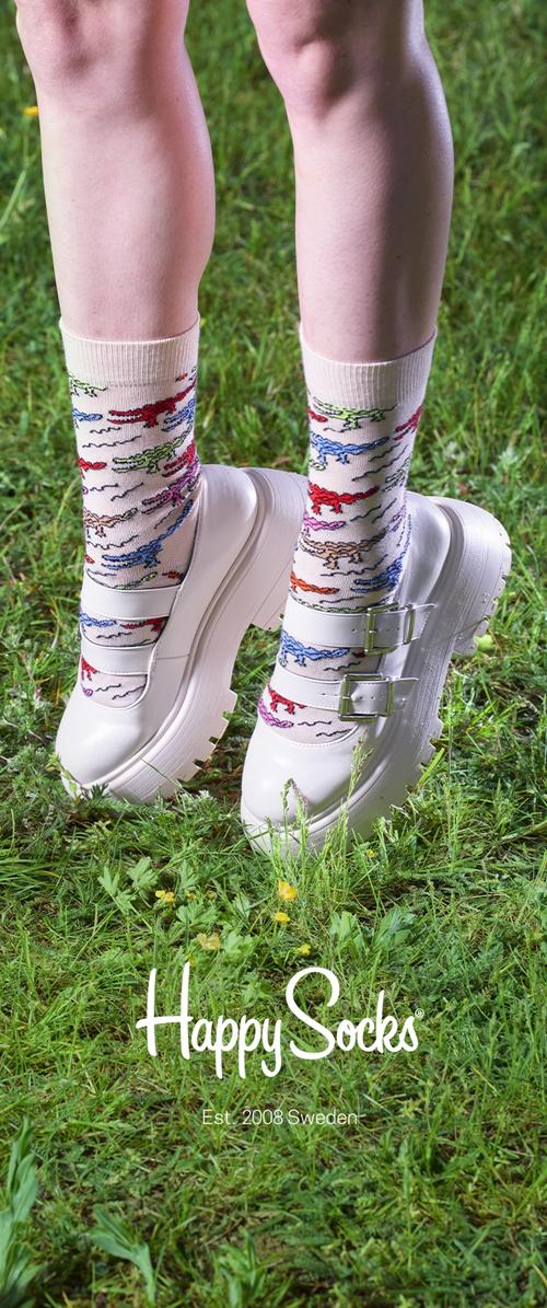 Weiße Plateau-Schuhe mit doppelten Schnallenriemen, getragen mit knielangen Socken, die bunte Dinosaurier-Muster auf einem grasbewachsenen Hintergrund zeigen.