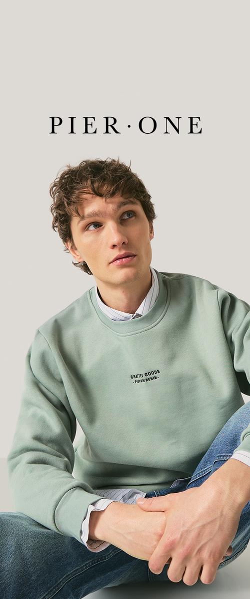 Joven con cabello rizado que lleva una sudadera verde claro y vaqueros, sentado con las manos entrelazadas, mirando ligeramente hacia arriba y a la izquierda.
