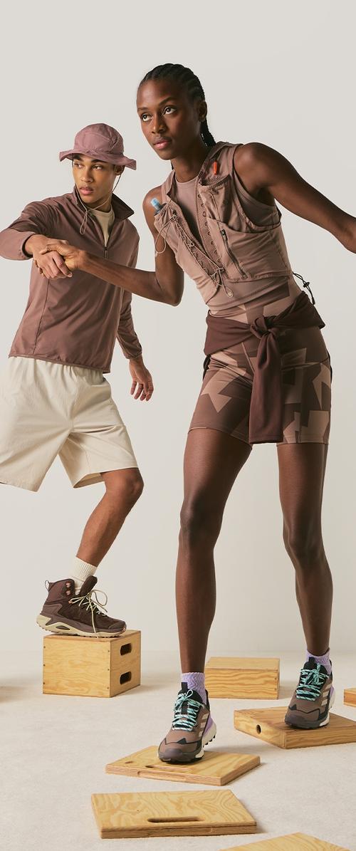 Deux athlètes vêtus de tenues de sport marron posant les pieds sur des plateformes en bois, l'un aidant l'autre à maintenir son équilibre en se tenant la main.
