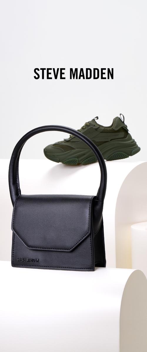 Bolso de mano de cuero sintético negro con un asa curvada y solapa frontal. Presenta el nombre de la marca grabado. Zapatillas deportivas verdes y gruesas en el fondo.