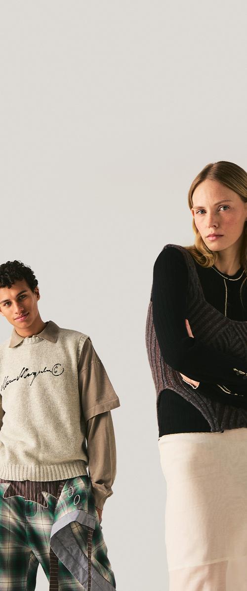 Joven hombre con ropa beige en capas y de cuadros está de pie, joven mujer con top negro y falda beige posa con los brazos cruzados, fondo gris neutro.
