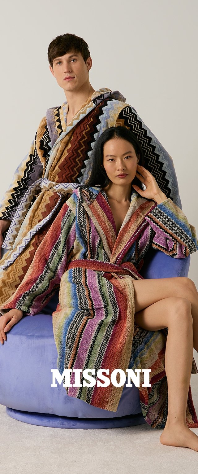 Hombre y mujer con albornoces coloridos con patrón en zigzag, mujer sentada en una silla morada, hombre de pie detrás de ella, fondo neutro.