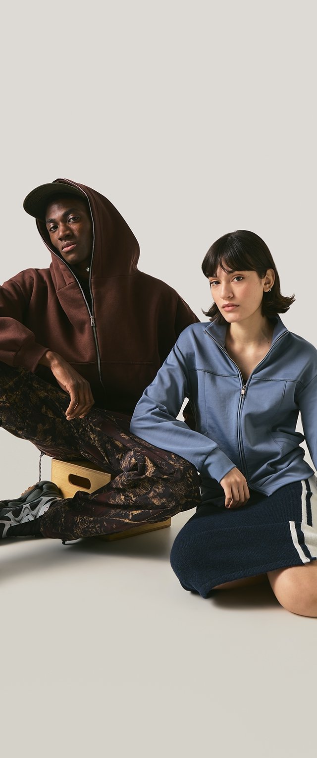 Due giovani seduti sul pavimento; uno indossa una felpa marrone con cappuccio, pantaloni a fantasia e scarpe da ginnastica; l'altro indossa una giacca blu con zip e una gonna blu.