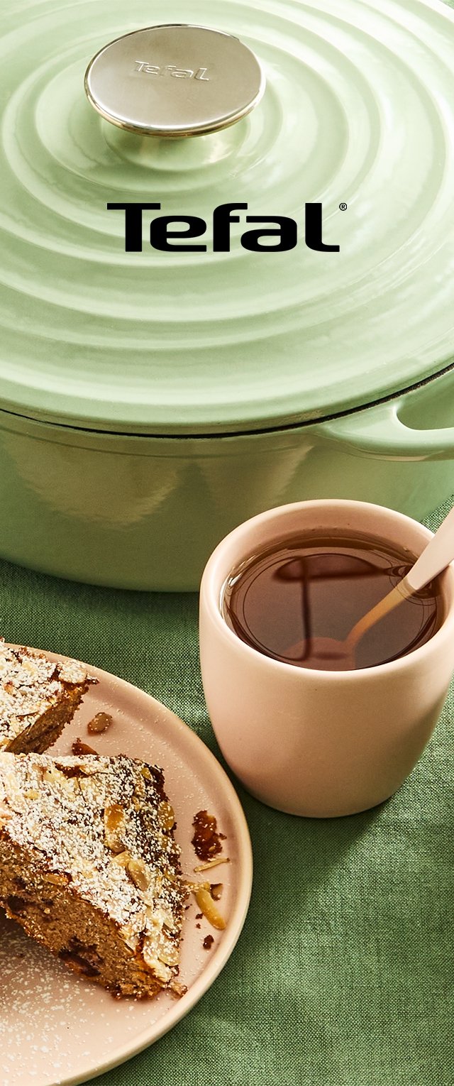 Grünes Tefal Kochgeschirr mit einem gerippten Deckel, eine hellrosa Tasse mit dunklem Inhalt und ein Stück Dessert auf einem beige-farbenen Teller auf einer grünen Oberfläche.