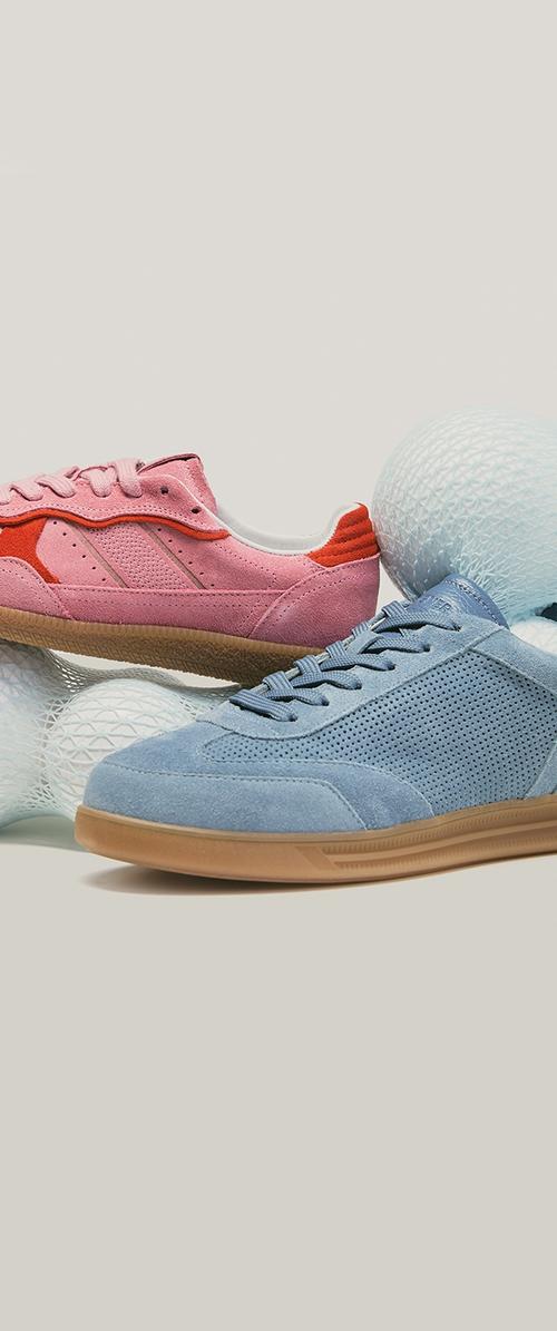 Paar Wildledersneaker, ein pinkes mit roten Akzenten und ein blaues mit hellbraunen Sohlen, ruhend auf weißen, strukturierten Kugeln vor einem neutralen Hintergrund.