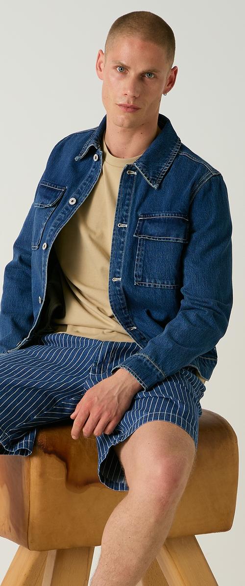 Jonge man met kort haar, gekleed in een beige shirt, een blauwe spijkerjas en blauwe shorts met fijne strepen, zittend op een houten kruk.