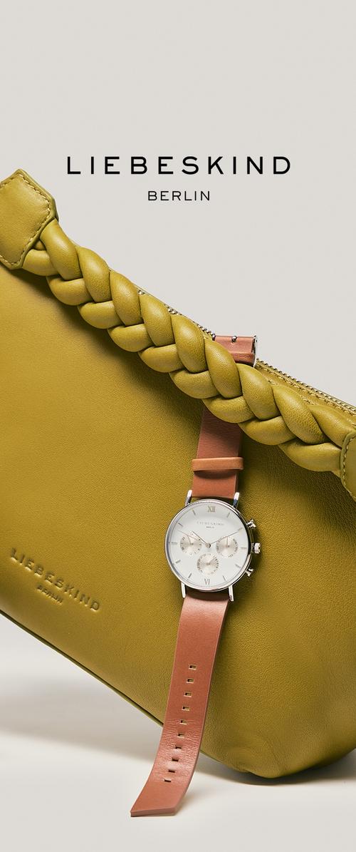 Bolso de cuero verde oliva con un tirador de cremallera trenzado y nombre de marca en relieve. Un reloj plateado con correa marrón se apoya contra el bolso.