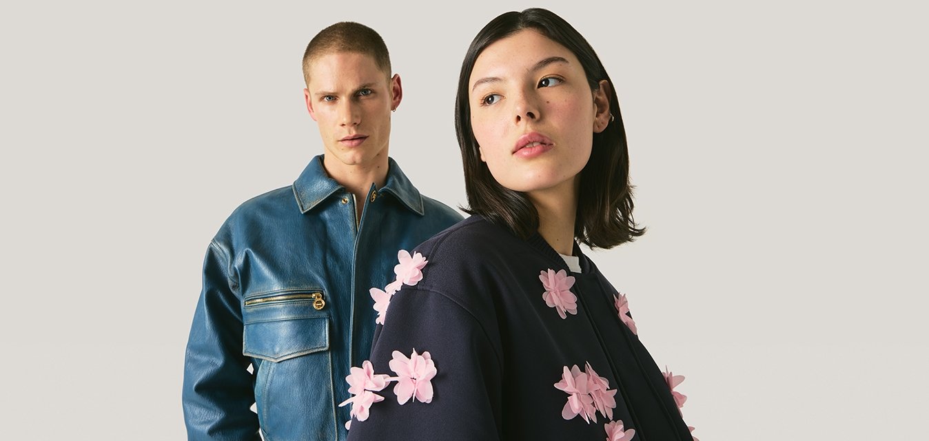 Dva modelové v módních bundách, jeden v modré kožené bundě, druhý v tmavě modré bundě s výraznými růžovými květinovými aplikacemi.