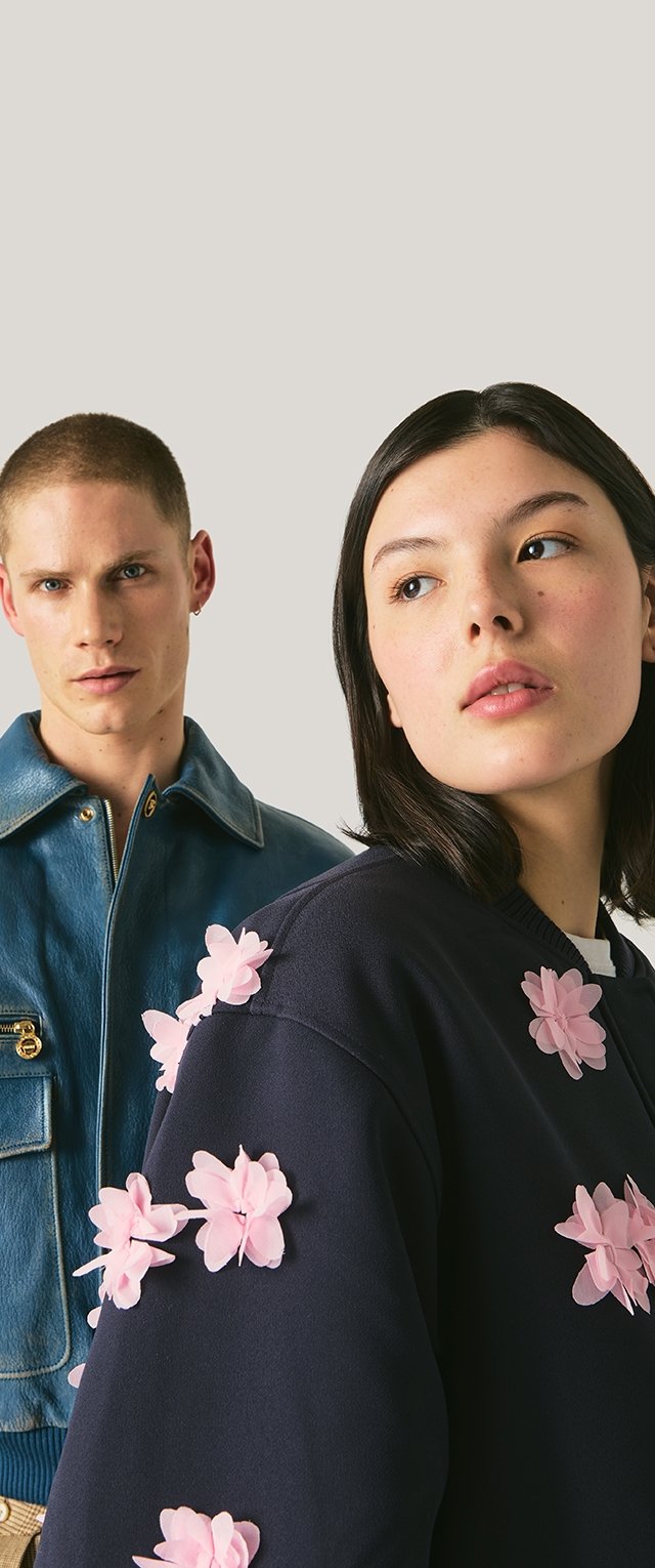 Joven con chaqueta de cuero azul y joven con suéter negro con apliques florales rosas, ambos mirando ligeramente hacia otro lado contra un fondo liso.