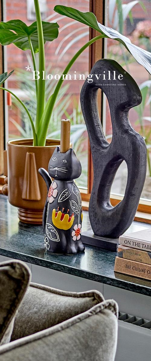 Bougeoir décoratif en forme de chat noir avec des motifs floraux, sculpture noire abstraite, plante en pot et livres empilés sur un rebord de fenêtre.