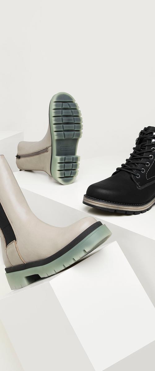 Three pairs of shoes: one beige ankle boot with black accents and green sole, and two black boots with textured leather and laces, featuring rubber soles.
