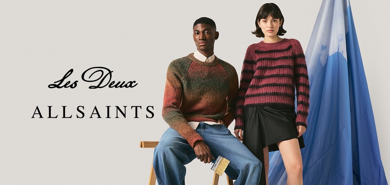 Dva modely, jeden sedící na stoličce v hnědém svetru a džínách, druhý stojící vedle modrého zavěšeného materiálu v pruhovaném svetru a černé sukni.