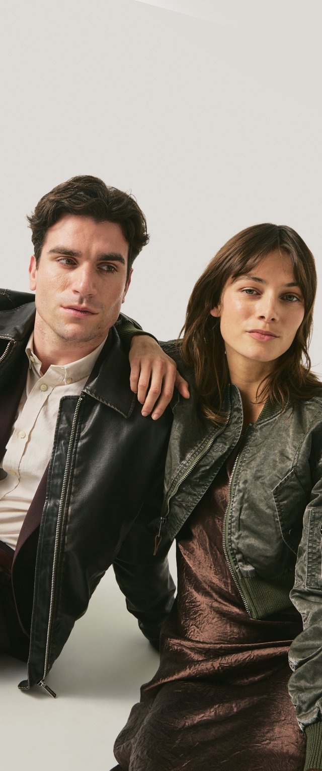 Un bărbat tânăr în geacă de piele neagră și cămașă albă stă lângă o tânără în geacă verde și rochie maro, brațul ei sprijinindu-se pe umărul lui.