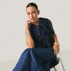 Žena sedící na židli, oblečená v námořnicky modré pletené vestě a odpovídající dlouhé sukni, opírající bradu o ruku, neutrální pozadí.