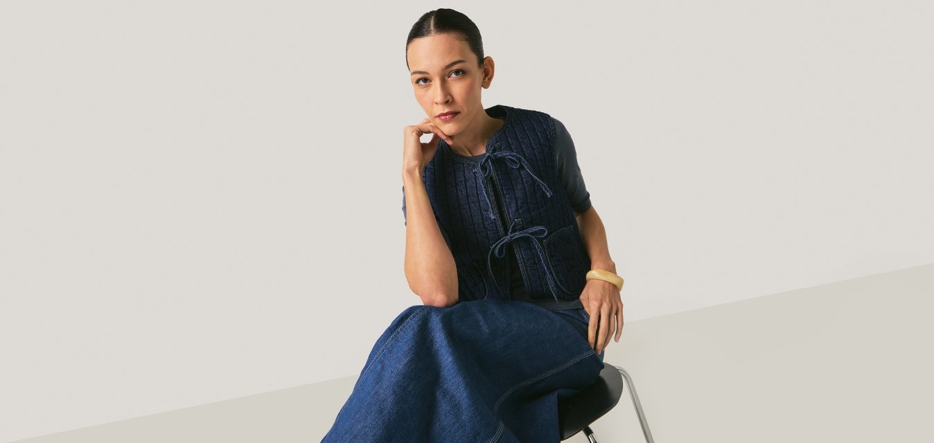 Žena sedící na židli, oblečená v námořnicky modré pletené vestě a odpovídající dlouhé sukni, opírající bradu o ruku, neutrální pozadí.