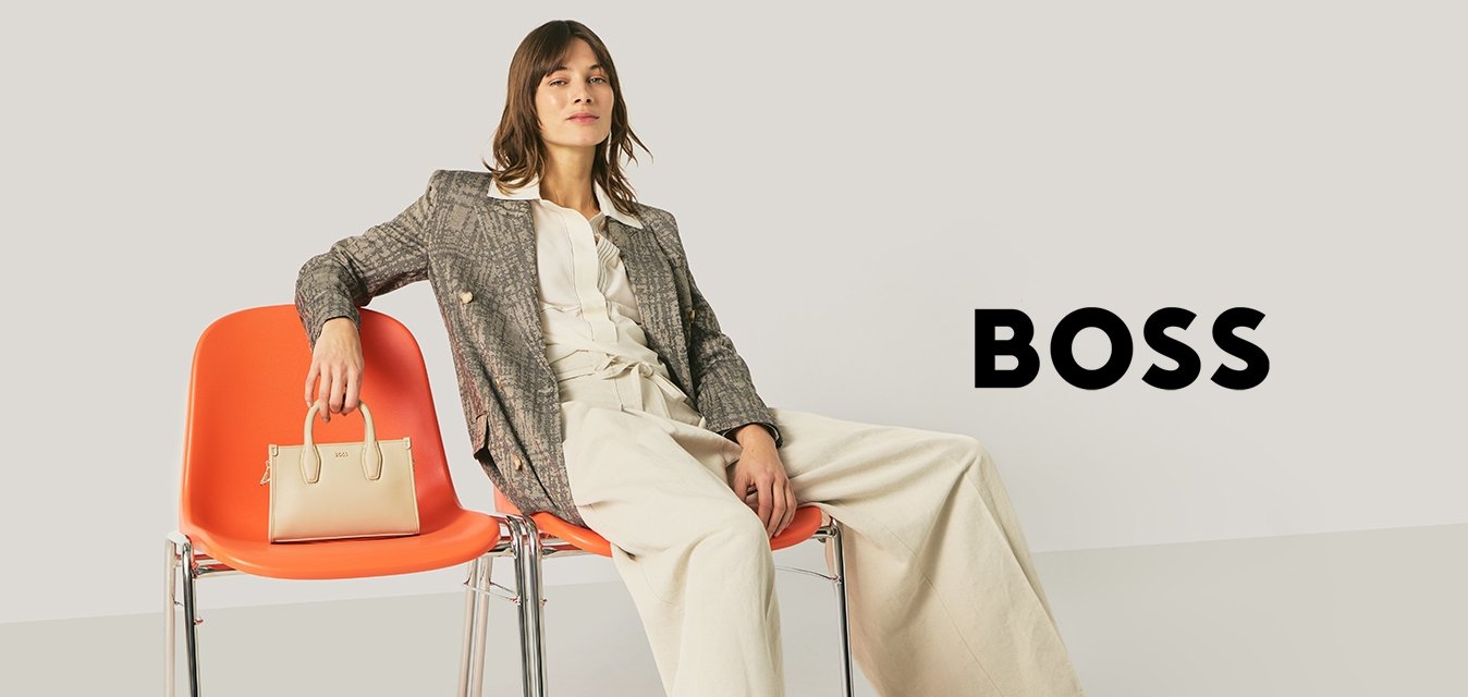 Žena v pracovním oblečení sedí uvolněně na dvou oranžových židlích, jednu židli drží béžovou kabelku, na pozadí je jednobarevná světlá stěna.