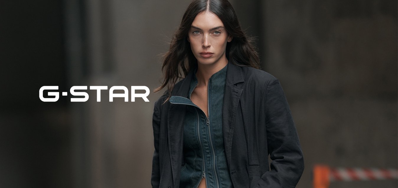 Žena s dlouhými tmavými vlasy, má na sobě rozepnutou tmavou bundu přes úzké džíny a tmavé sako, stojící ve městě.