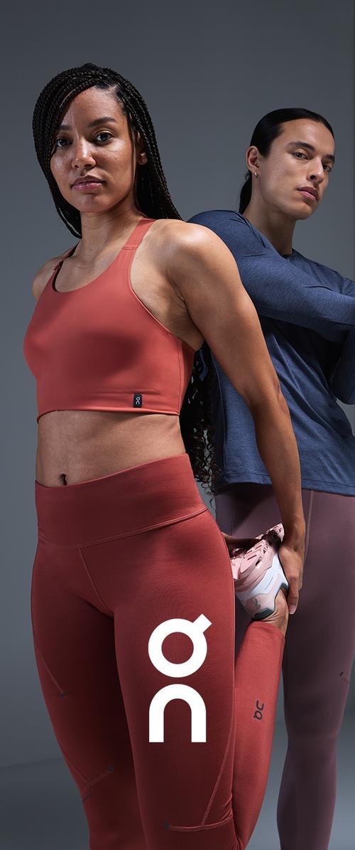Sostenedor deportivo rojo y leggings de cintura alta, ambos con una textura suave y acentos de logo. Top de manga larga gris y zapatilla rosa pálido en el fondo.